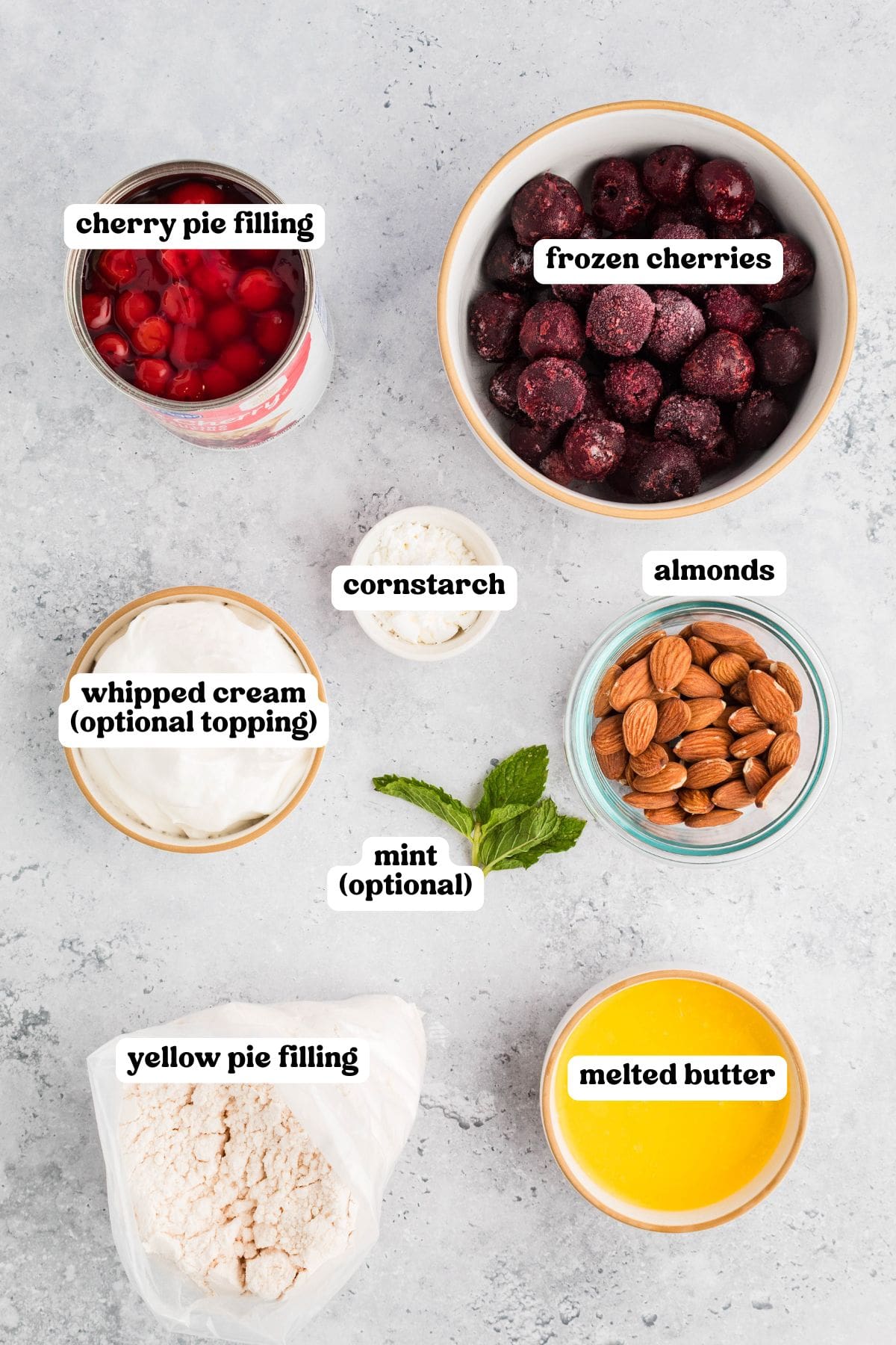 Ingredients for cherry cobbler displayed on a gray surface, each labeled: cherry pie filling, frozen cherries, cornstarch, almonds, whipped cream (optional topping), mint (optional), yellow pie filling mix, and melted butter.