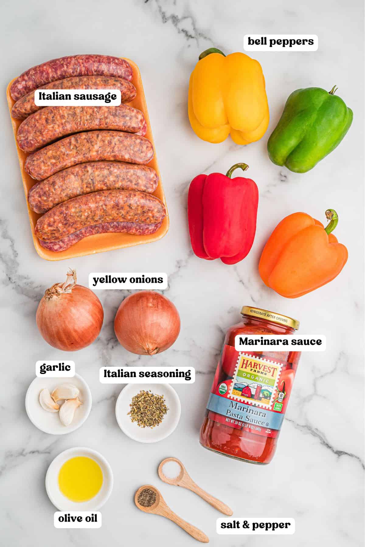 Ingredients for sausage and peppers laid out on a marble surface with labels: a pack of raw Italian sausages, yellow, green, red, and orange bell peppers, two yellow onions, garlic cloves, Italian seasoning, a jar of marinara sauce, a small dish of olive oil, and wooden spoons holding salt and pepper.