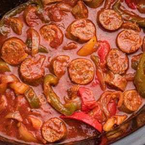 Overhead image showing sliced sausage, green, red, and yellow bell peppers, and onions simmered in red marinara sauce inside a crockpot.