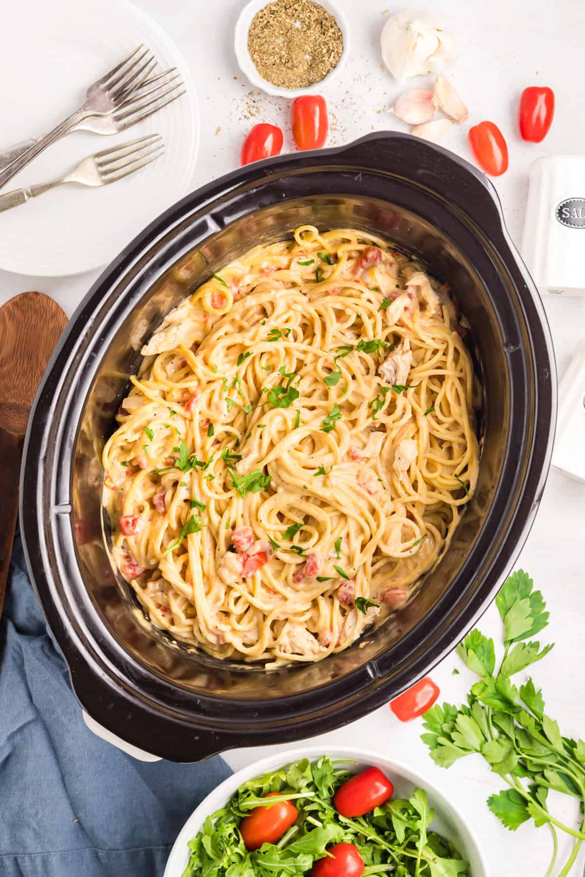 Overhead view of a slow cooker filled with creamy chicken spaghetti with rotel. Around it are forks, a wooden spoon, garlic cloves, tomatoes, Italian seasoning, cream cheese, salt container, parsley, and a small bowl of greens.