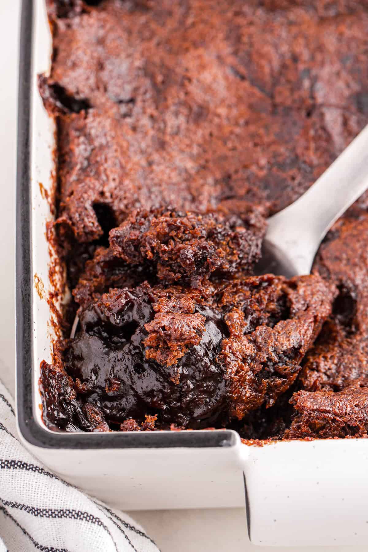 Close-up of chocolate pudding cake in a white rectangular baking dish with a spoon scooping out a portion. The cake has a dark, shiny chocolate sauce layer and a soft, slightly crumbly top.