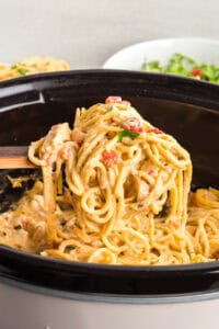 Side view of a wooden spoon lifting creamy chicken spaghetti with visible diced tomatoes and chunks of chicken from slow cooker.