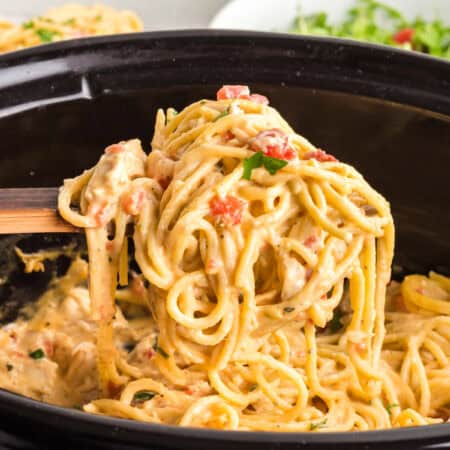 Side view of a wooden spoon lifting creamy chicken spaghetti with visible diced tomatoes and chunks of chicken from slow cooker.
