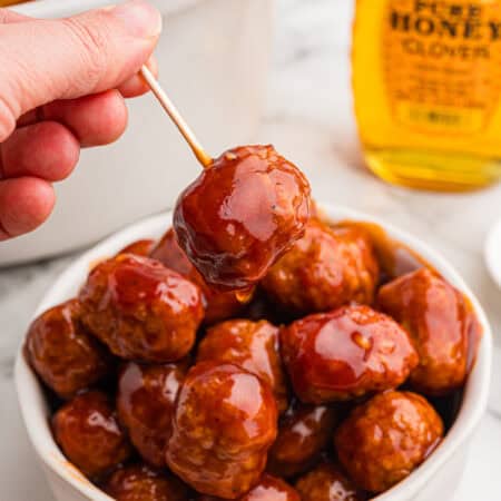 A hand holds a glazed honey BBQ meatball on a toothpick above a bowl of meatballs.