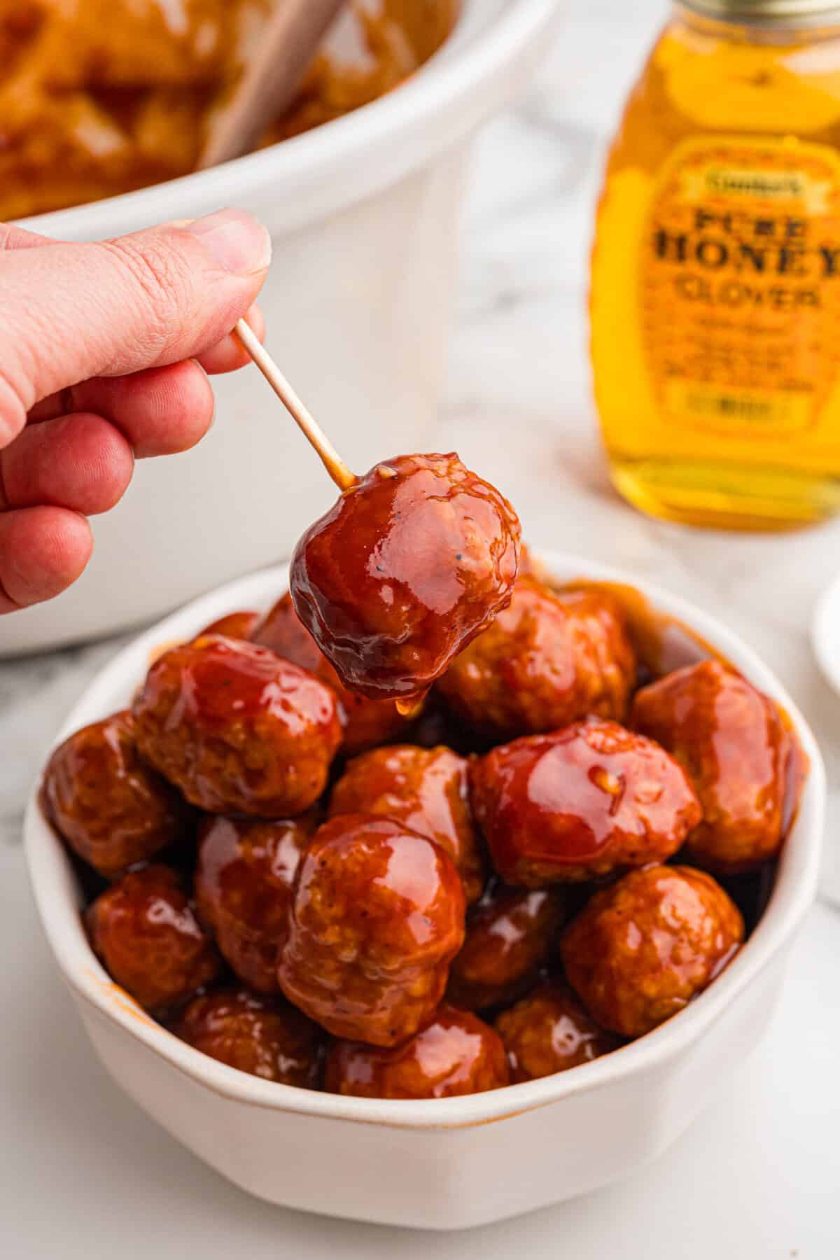 A hand holds a glazed honey BBQ meatball on a toothpick above a bowl of meatballs.