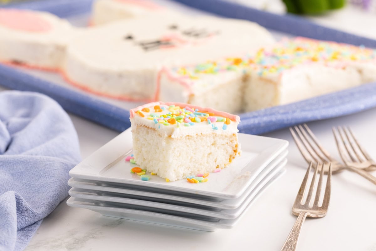 Slice of frosted Easter bunny cake on stacked white plates with sprinkles.