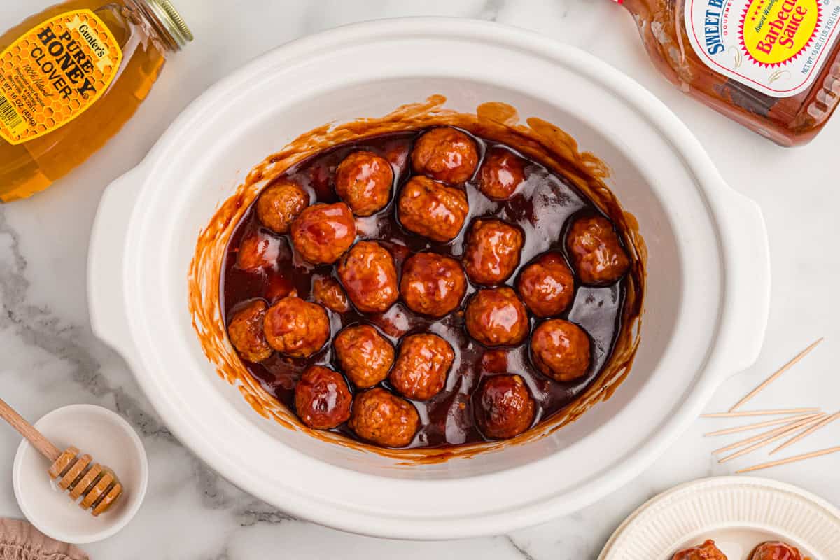 Overhead view of saucy bbq meatballs in a slow cooker with honey and bbq sauce nearby.