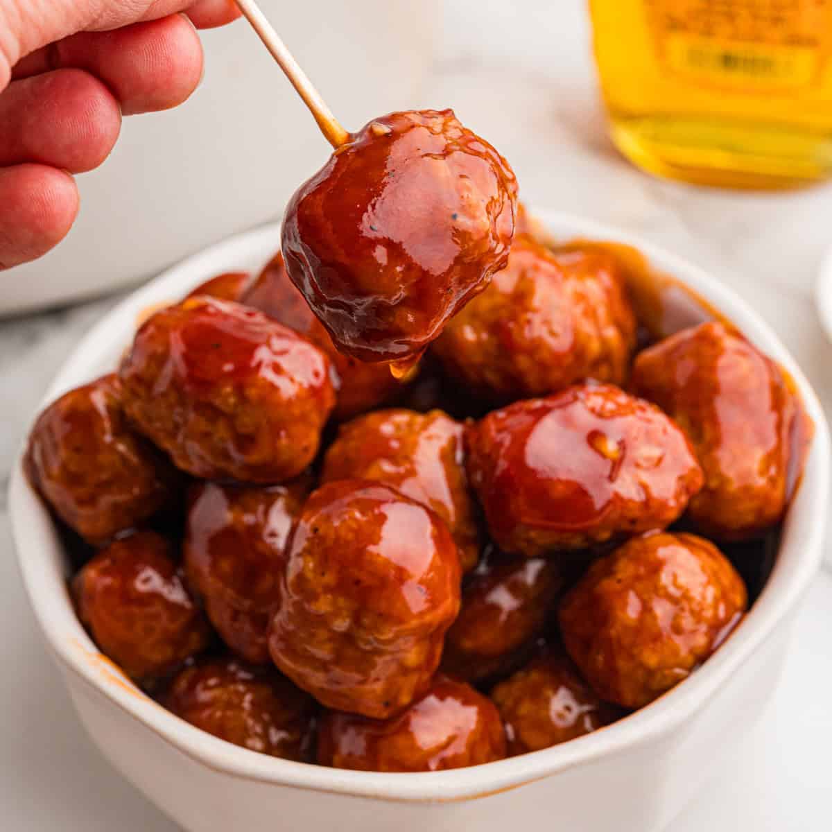 A hand holds a glazed honey BBQ meatball on a toothpick above a bowl of meatballs.