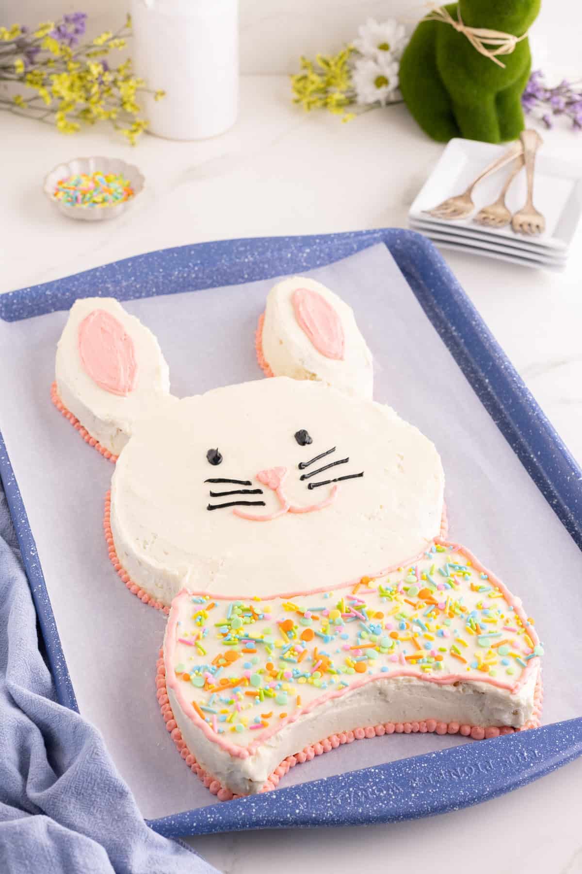 Bunny-shaped cake with white frosting and pink inner ears, black eyes and whiskers, and a pink nose and mouth. The lower half is decorated like a bow tie with pastel sprinkles and outlined in pink piping.
