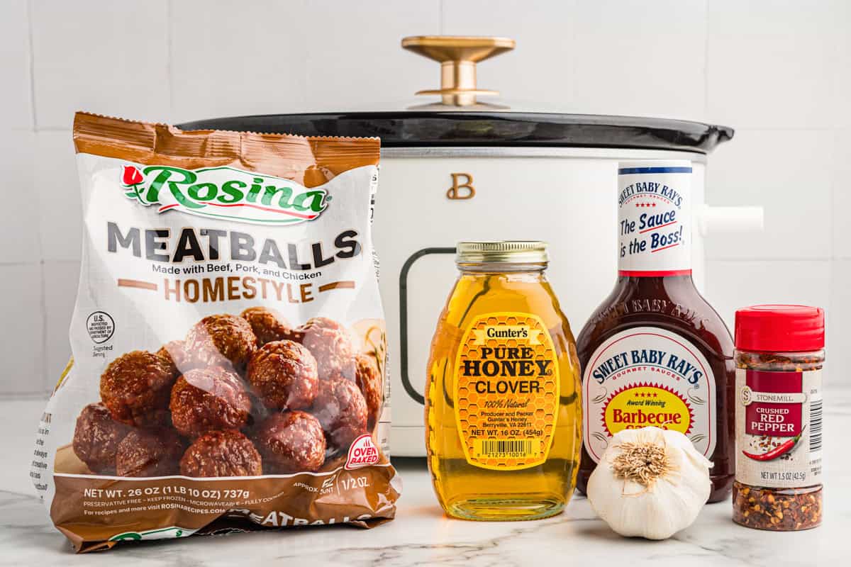 A package of frozen Rosina homestyle meatballs, a jar of honey, Sweet Baby Ray’s barbecue sauce, garlic, crushed red pepper, and a slow cooker are arranged on a counter.