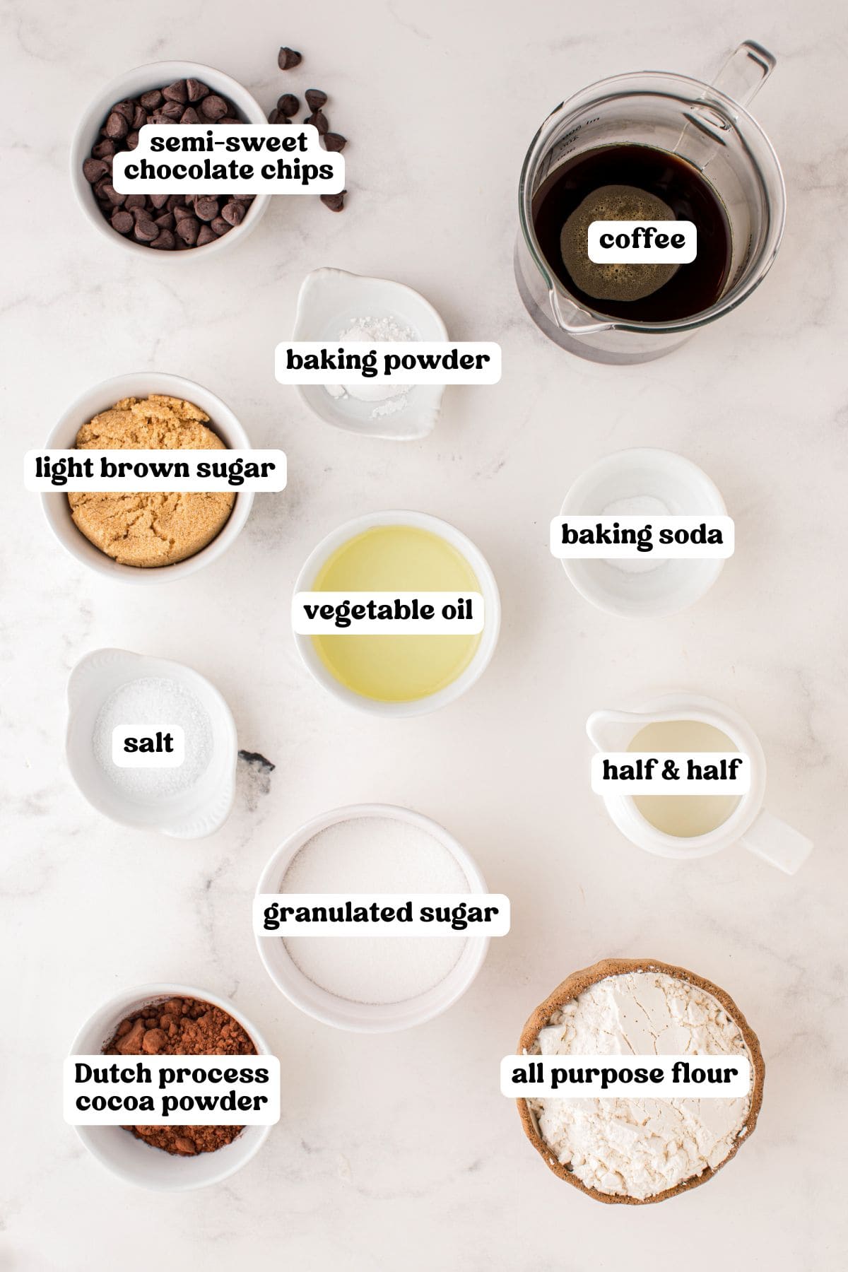 verhead view of baking ingredients arranged in white bowls on a light surface, each labeled: semi-sweet chocolate chips, coffee in a glass measuring cup, baking powder, light brown sugar, baking soda, vegetable oil, salt, half and half, granulated sugar, Dutch process cocoa powder, and all purpose flour.