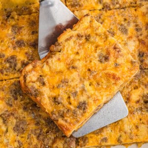 Close-up of a golden-brown sausage breakfast casserole with melted cheddar and crumbled sausage on top. A square piece is being lifted from the pan with a metal spatula, showing the layered interior.