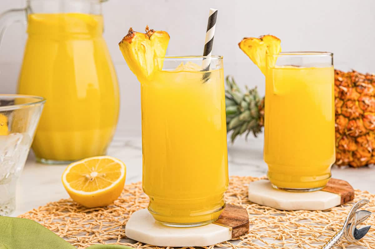 Two glasses of pineapple lemonade with pineapple garnishes, with a pitcher and halved lemon nearby.