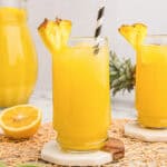 Glass of pineapple lemonade punch with ice, a pineapple wedge garnish, and a striped paper straw, with a second glass and pitcher in the background.