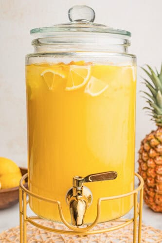 Glass drink dispenser filled with pineapple lemonade, ice, and lemon slices on a gold stand.