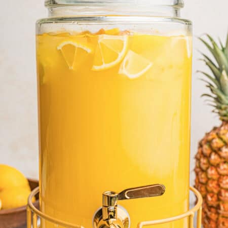 Glass drink dispenser filled with pineapple lemonade, ice, and lemon slices on a gold stand.