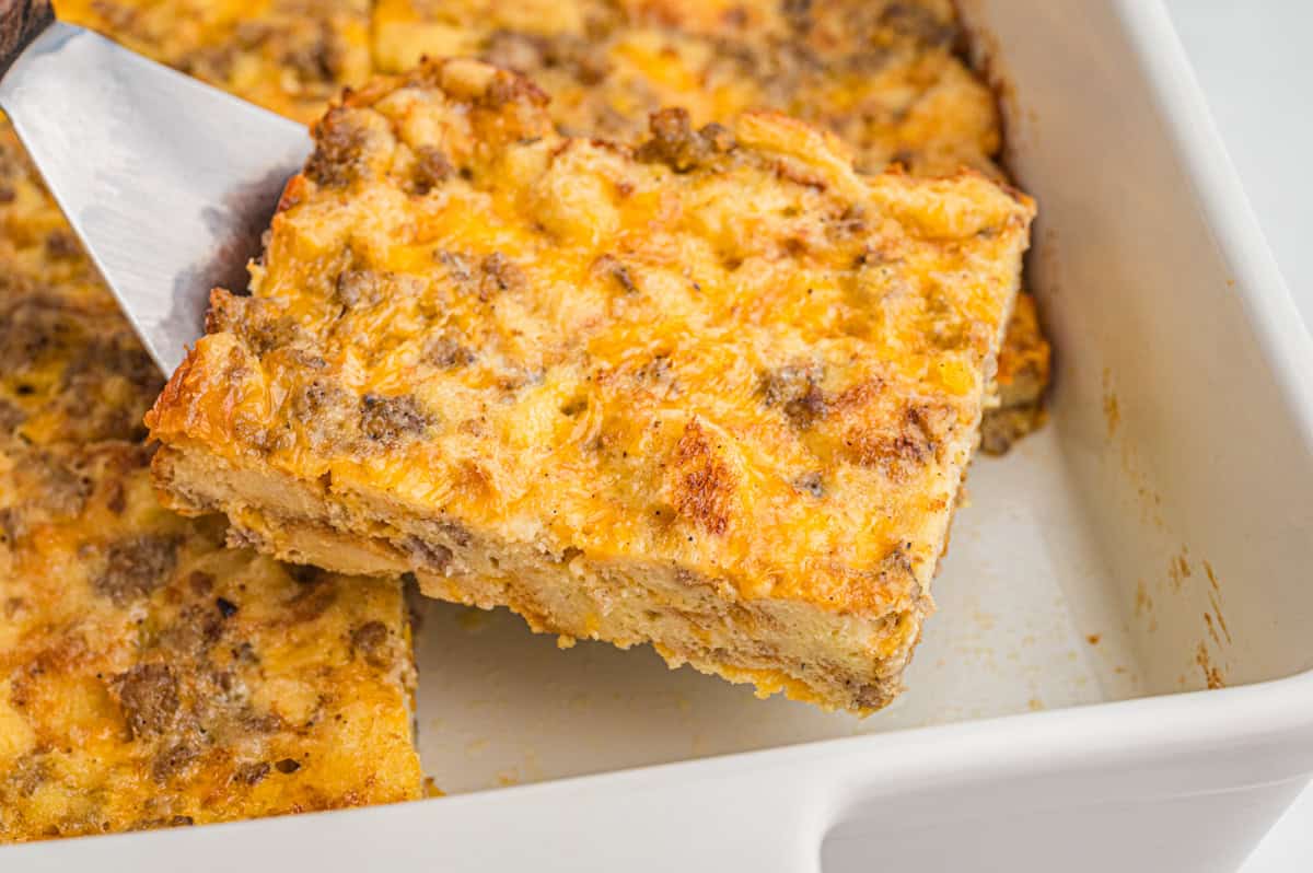 Close-up of an overnight breakfast casserole serving being lifted from a white baking dish with a metal spatula. The casserole shows layers of bread, egg, sausage, and melted cheddar with a golden-brown top.