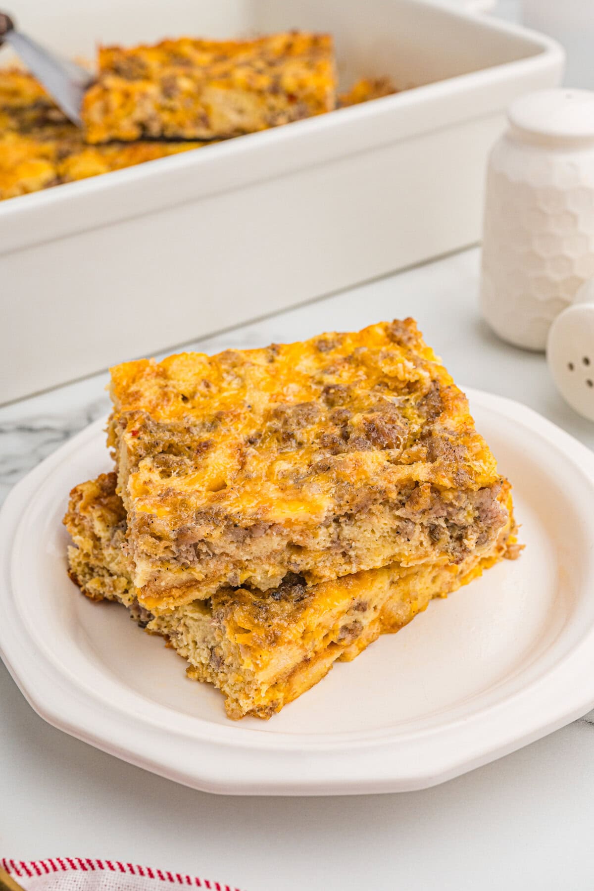 Two square slices of sausage and egg breakfast casserole stacked on a white plate, with the baking dish in the background.