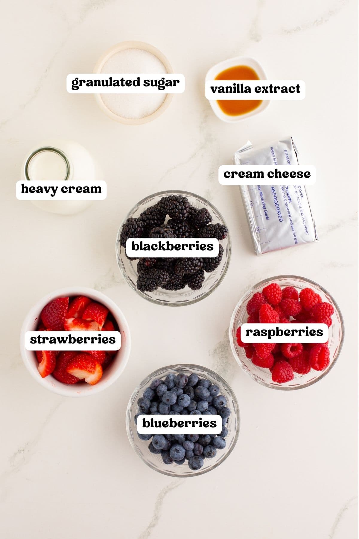 Labeled ingredients including bowls of strawberries, blueberries, raspberries, and blackberries, a block of cream cheese, a container of heavy cream, a bowl of granulated sugar, and a small dish of vanilla extract.