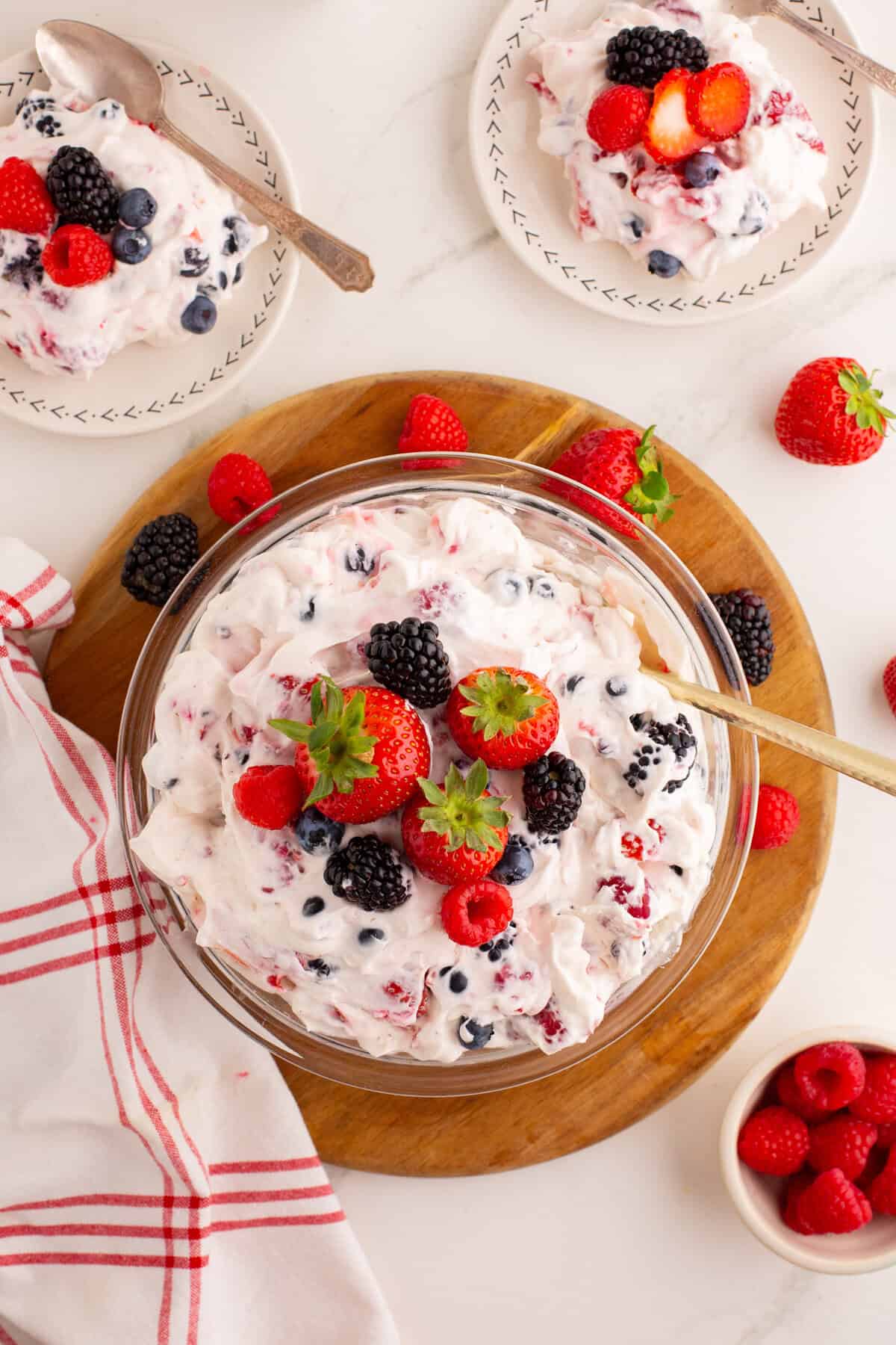 Glass bowl filled with mixed berry cheesecake salad. Fresh strawberries, raspberries, and blackberries surround the bowl, with a spoon resting inside and small plates of salad nearby.
