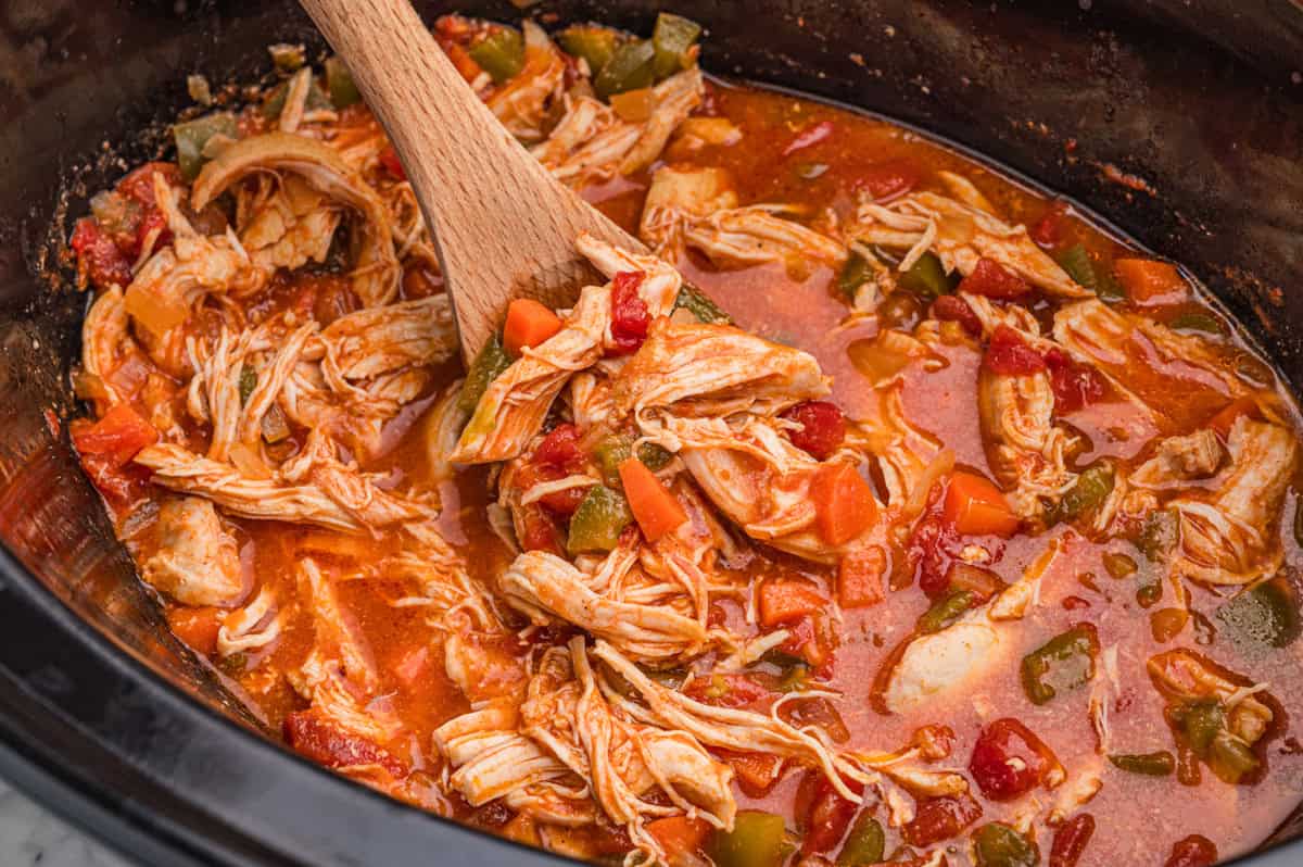 Spoon lifting crock pot chicken paprika coated in a red sauce with visible diced tomatoes from a slow cooker.
