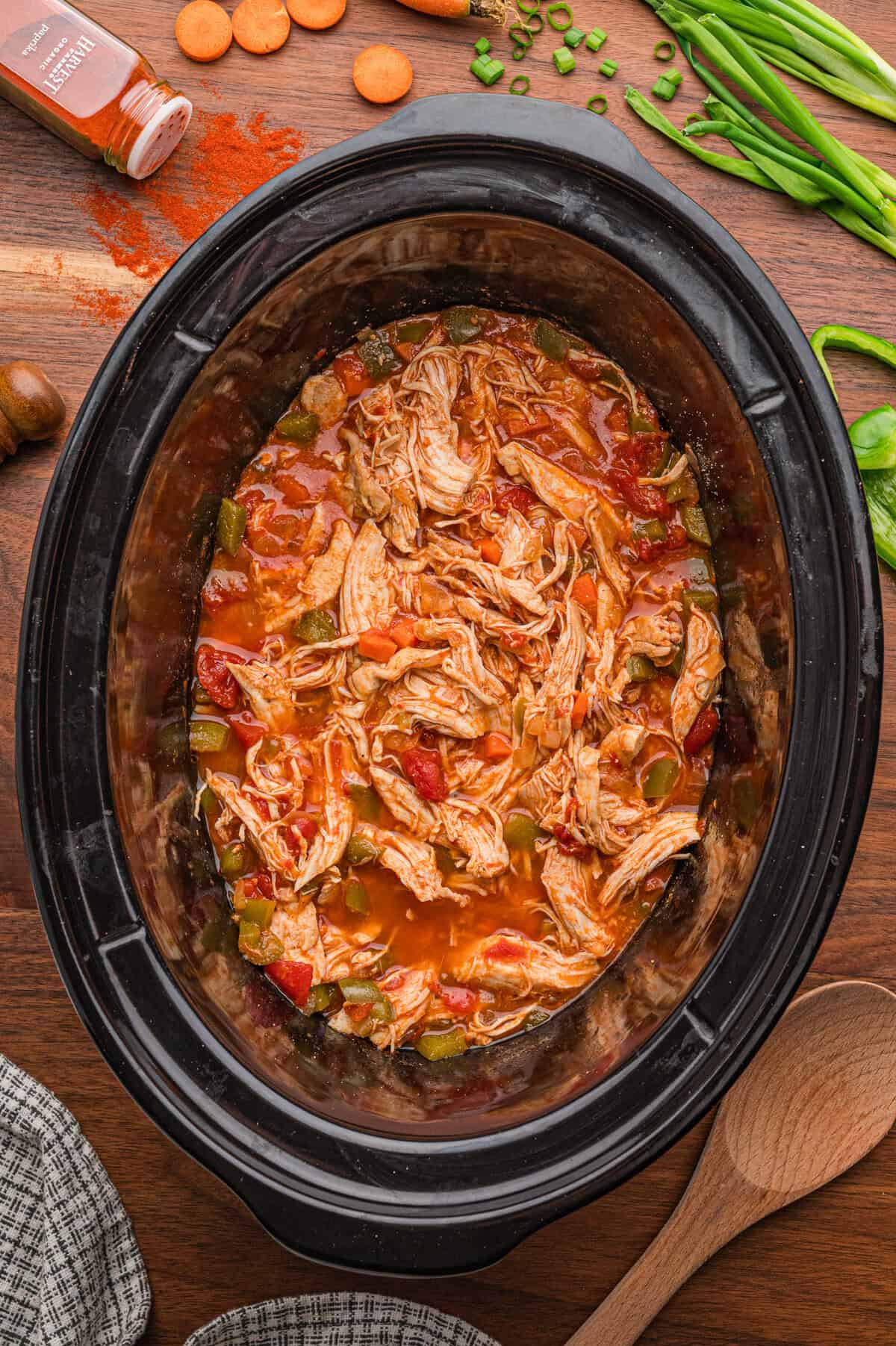 Slow Cooked Chicken Paprika surrounded by sliced carrots, green onions, paprika seasoning, and a wooden spoon on a wooden table.