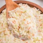 Close-up of a wooden spoon lifting a heaping scoop of finely shredded coleslaw from a wooden bowl.