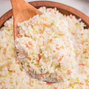 Close-up of a wooden spoon lifting a heaping scoop of finely shredded coleslaw from a wooden bowl.