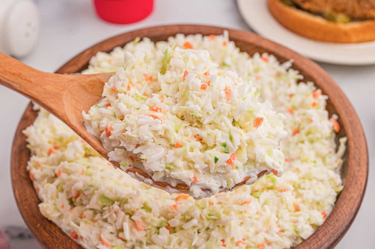 Wooden bowl of coleslaw with a wooden spoon, a chicken sandwich on a plate in the background.
