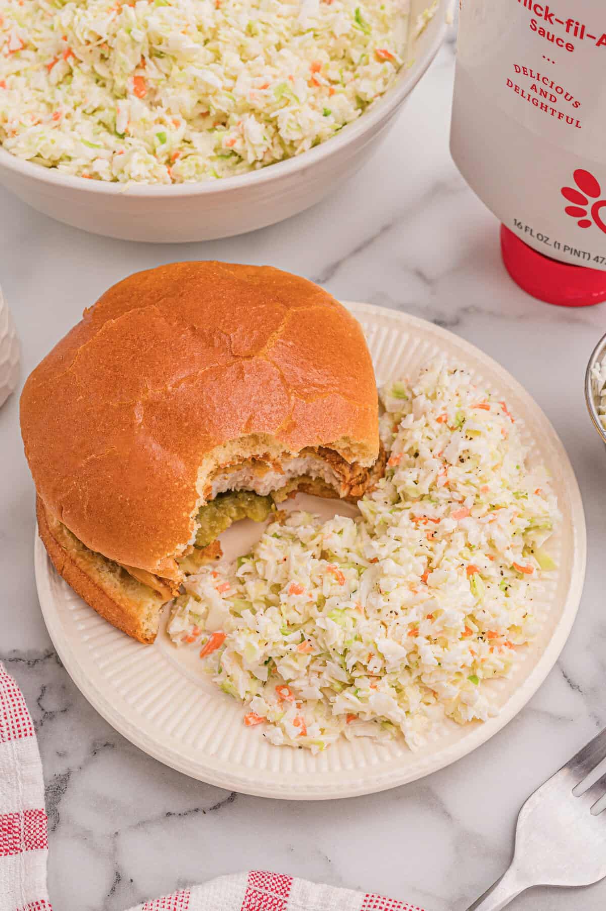 Chicken sandwich plated with coleslaw on the side, a bottle of Chick-fil-A sauce in the background.
