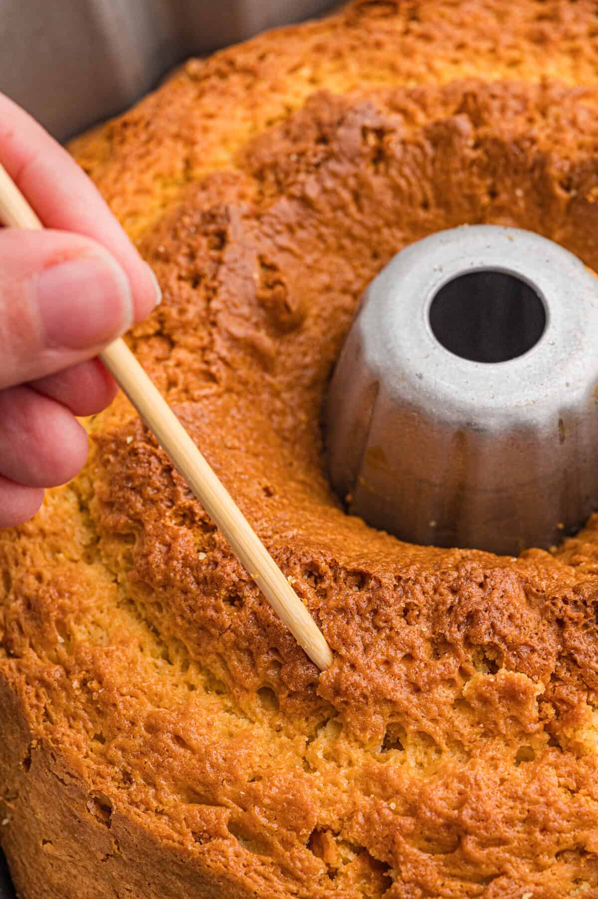 A hand inserting a wooden toothpick into a baked rum cake in a bundt pan to test doneness.