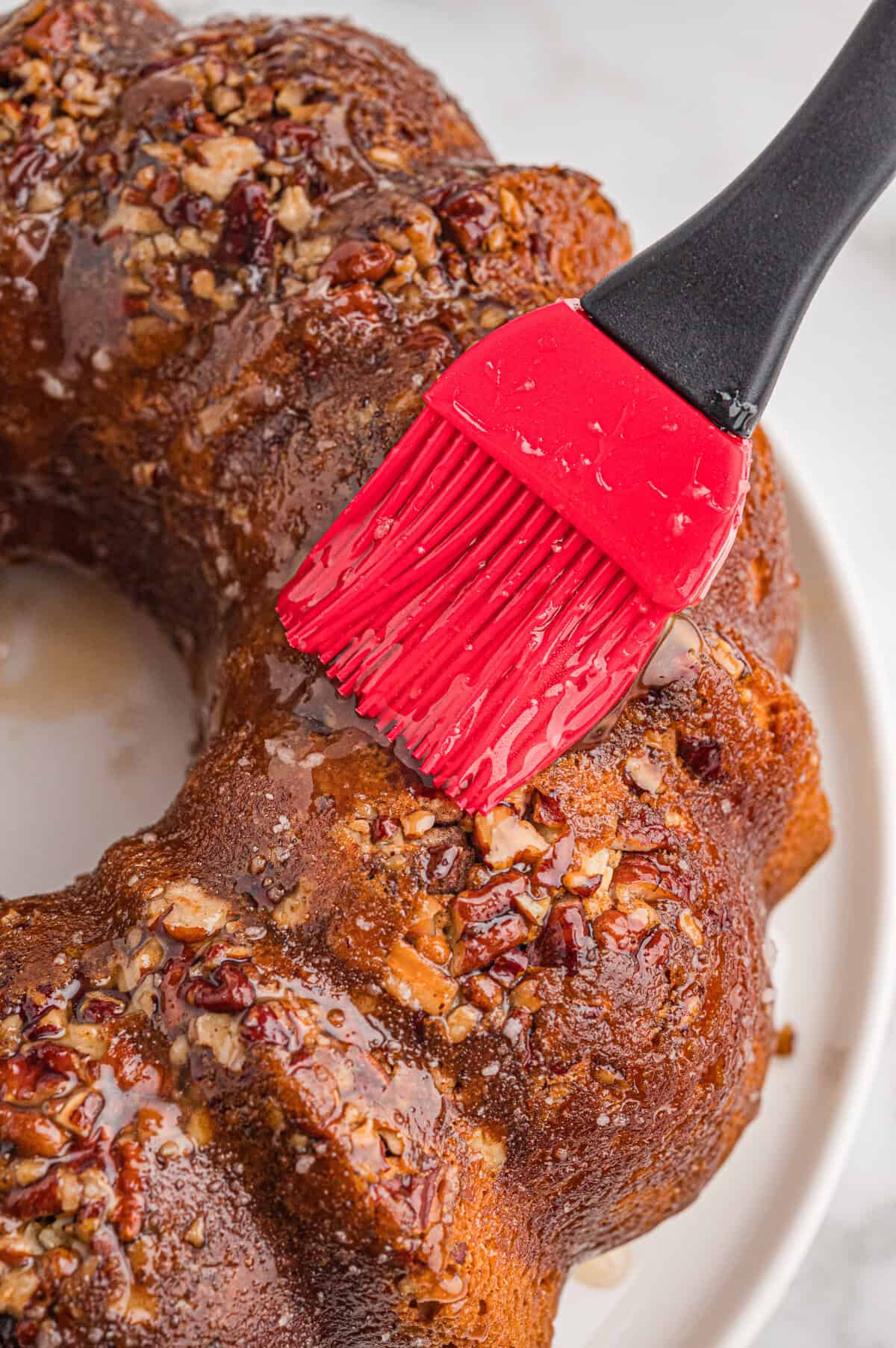 Red silicone pastry brush brushing rum glaze onto a baked bundt cake topped with pecans.
