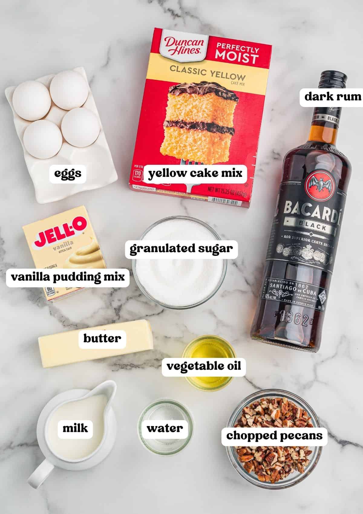 Rum cake ingredients on marble: eggs, yellow cake mix, vanilla pudding, sugar, butter, oil, milk, water, pecans, dark rum.