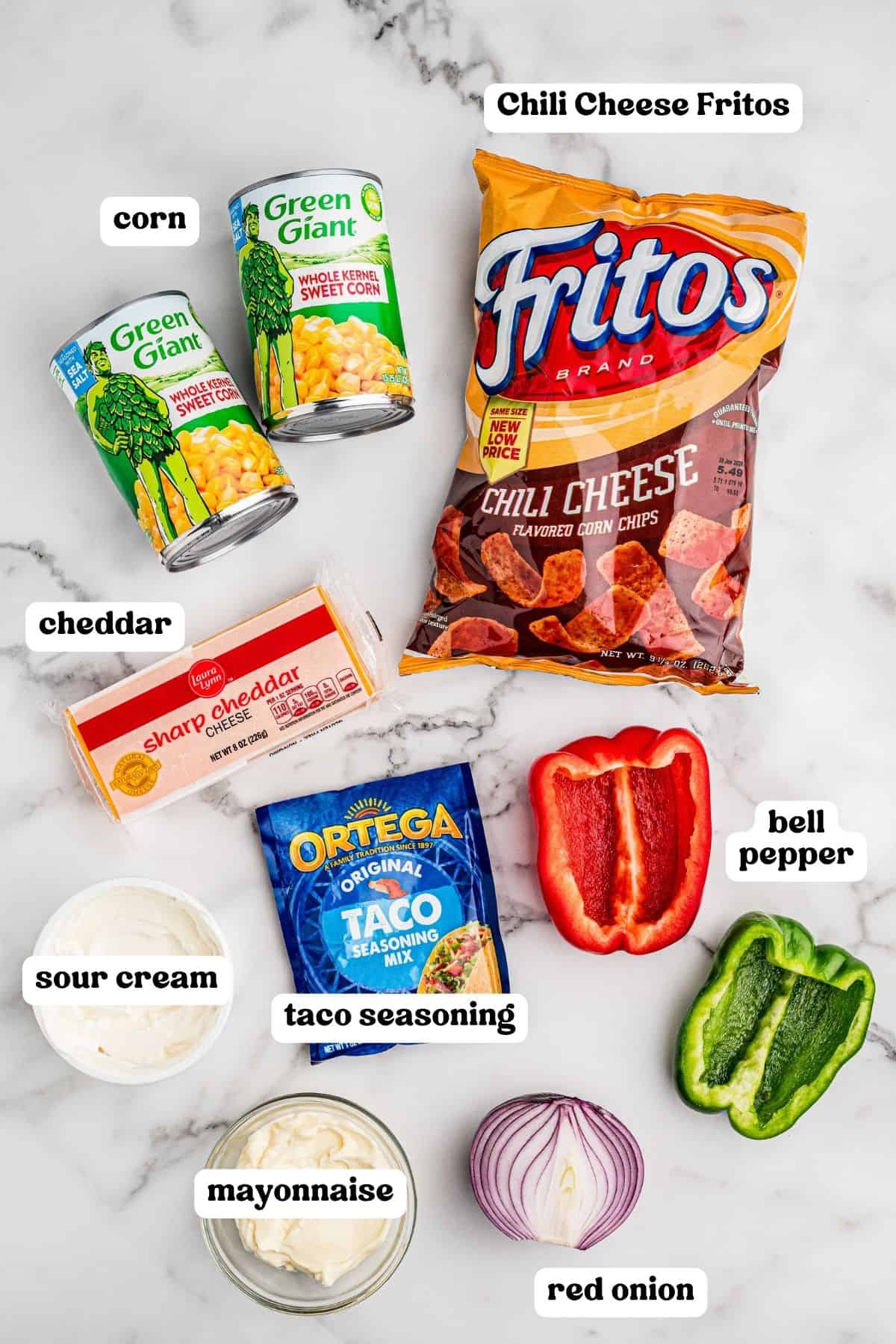 Ingredients on a marble counter: two cans of Green Giant whole kernel corn, a bag of Chili Cheese Fritos, a block of sharp cheddar, sour cream, mayonnaise, an Ortega taco seasoning packet, half a red bell pepper, half a green bell pepper, and half a red onion.