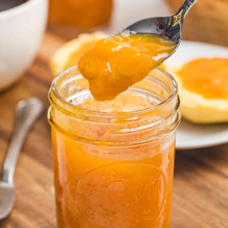 A spoon lifting glossy apricot jam from a mason jar with biscuit halves and apricots in the background.