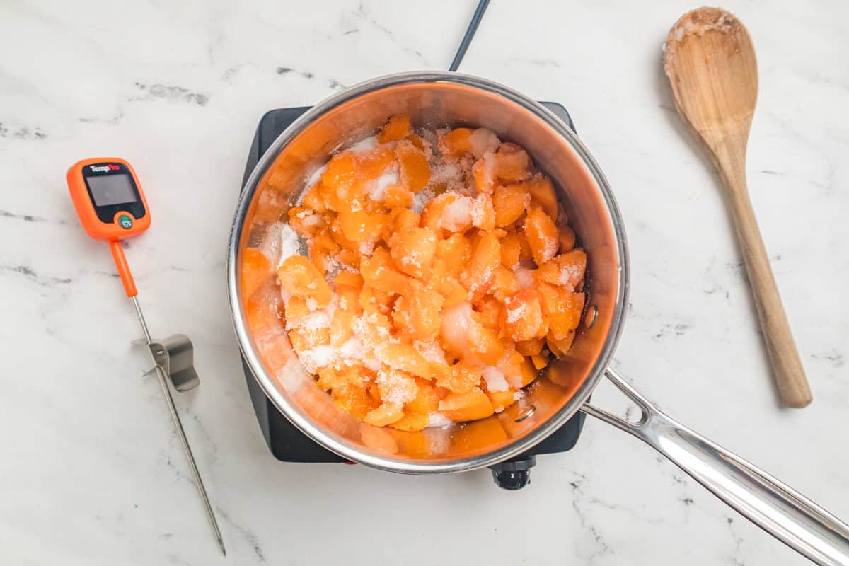 Diced apricots in a saucepan sprinkled with granulated sugar with a thermometer and a wooden spoon nearby.