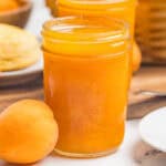 A close-up of a mason jar filled with apricot jam beside a single whole apricot on a marble counter.
