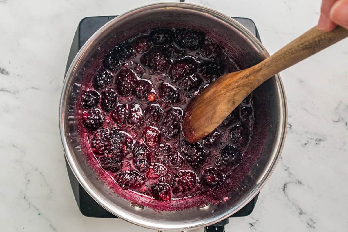 Whole blackberries breaking down in a saucepan with a wooden spoon stirring the dark juice.