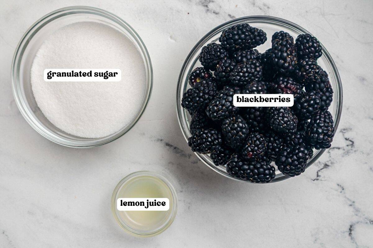 Ingredients for blackberry jam labeled on a marble counter: granulated sugar, fresh blackberries, and lemon juice.