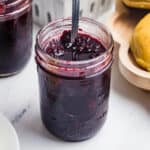 A close-up square shot of an open mason jar of blackberry jam with a spoon stirring the surface.