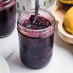 A close-up square shot of an open mason jar of blackberry jam with a spoon stirring the surface.