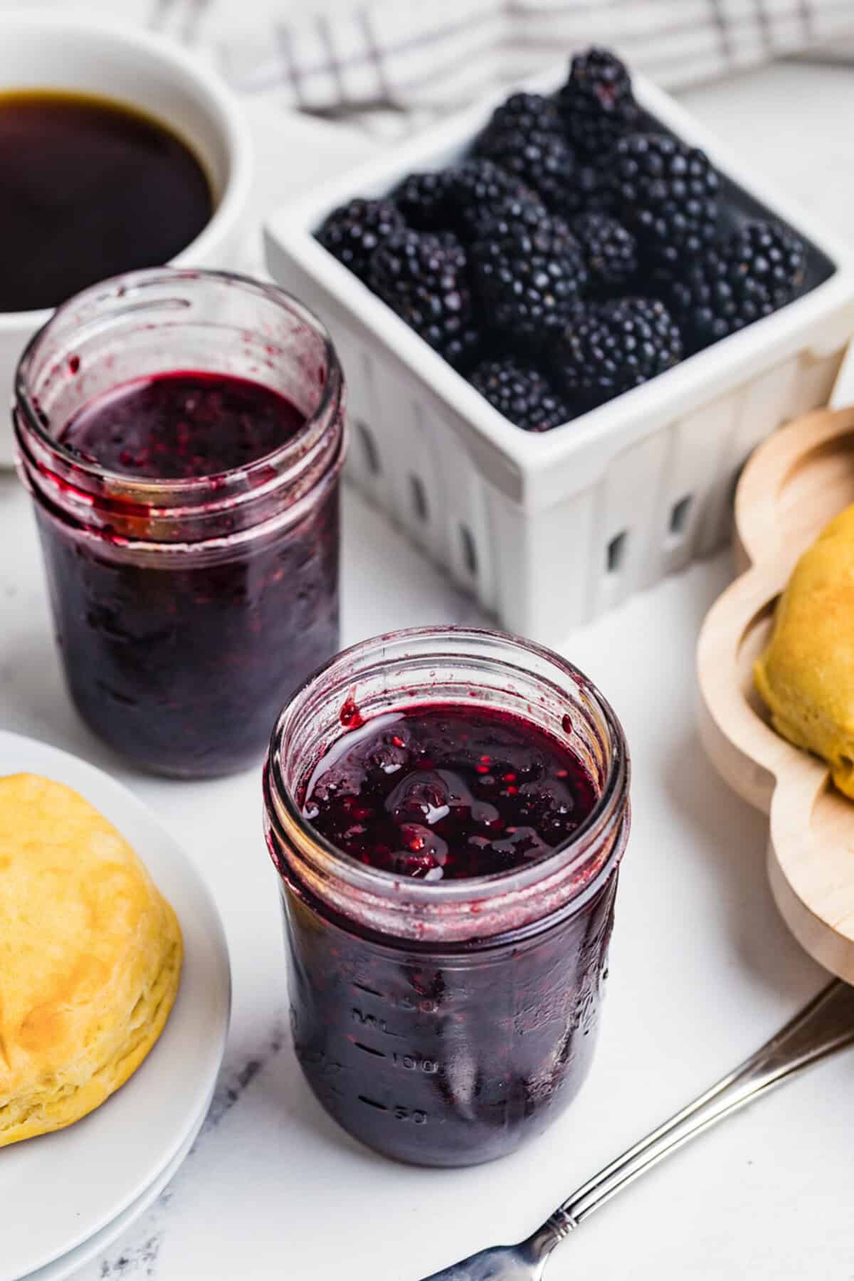 Two open jars of homemade blackberry jam on a counter with a basket of fresh blackberries, a coffee cup, and a biscuit.