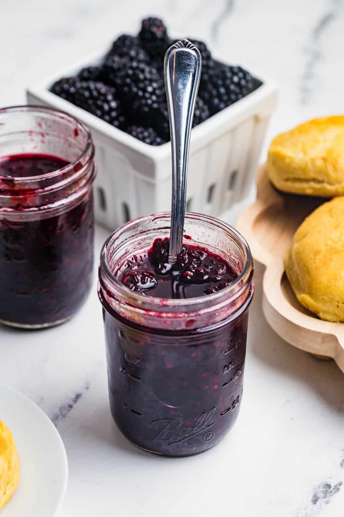 A tall open mason jar of blackberry jam with a spoon resting in it and a basket of fresh blackberries in the background.