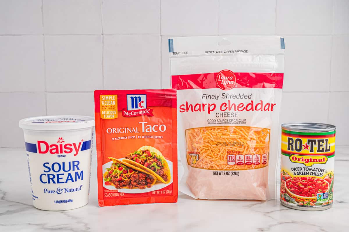 Packaged ingredients on a marble counter: Daisy sour cream, taco seasoning, shredded sharp cheddar, and a can of Rotel.