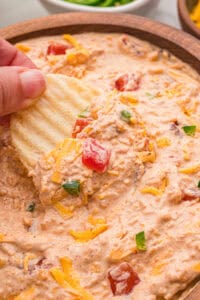 A hand scooping creamy dip from a wooden bowl with a ruffled potato chip.
