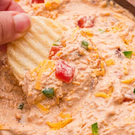 A hand scooping creamy dip from a wooden bowl with a ruffled potato chip.