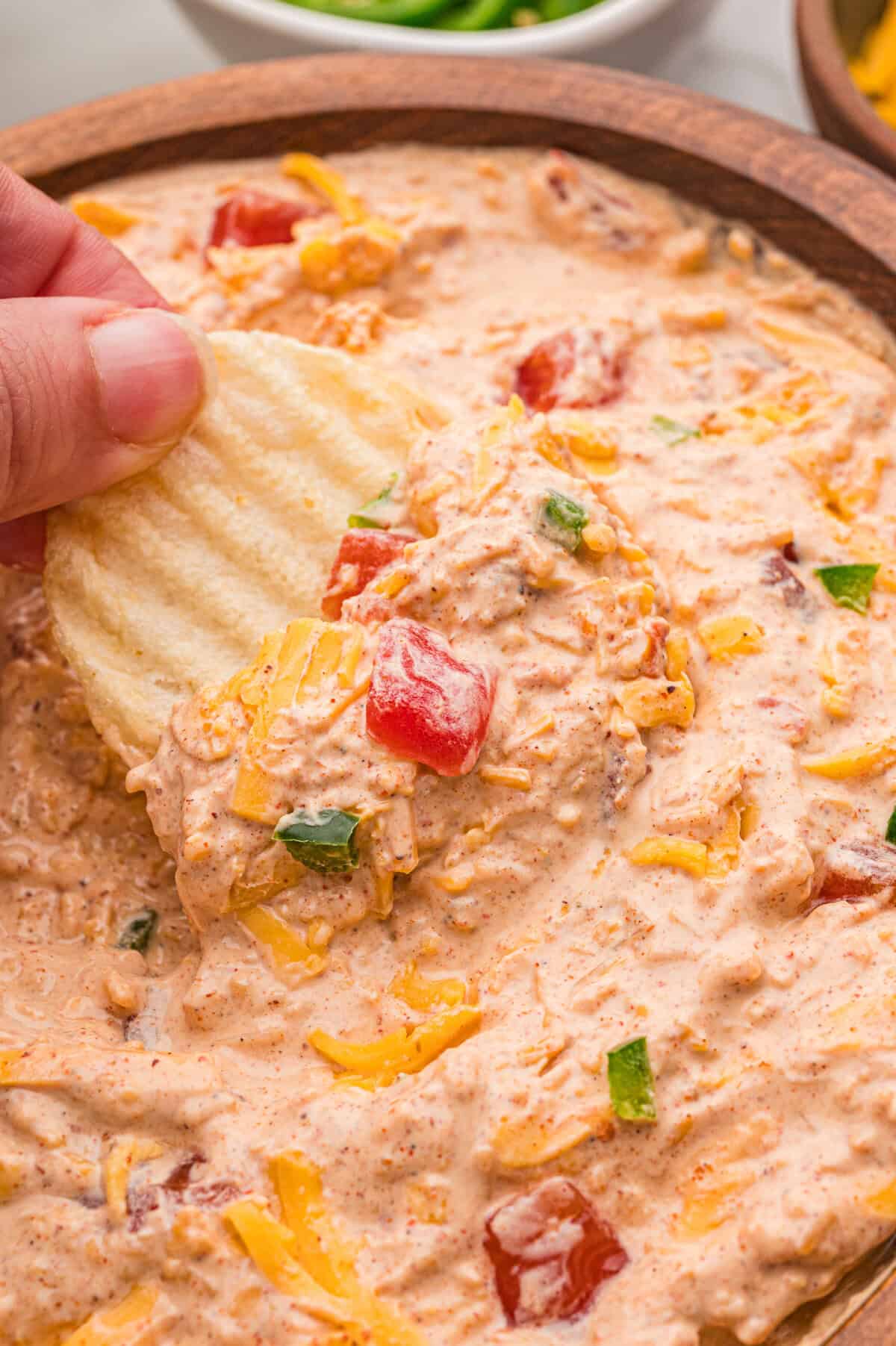 A hand scooping creamy dip from a wooden bowl with a ruffled potato chip.