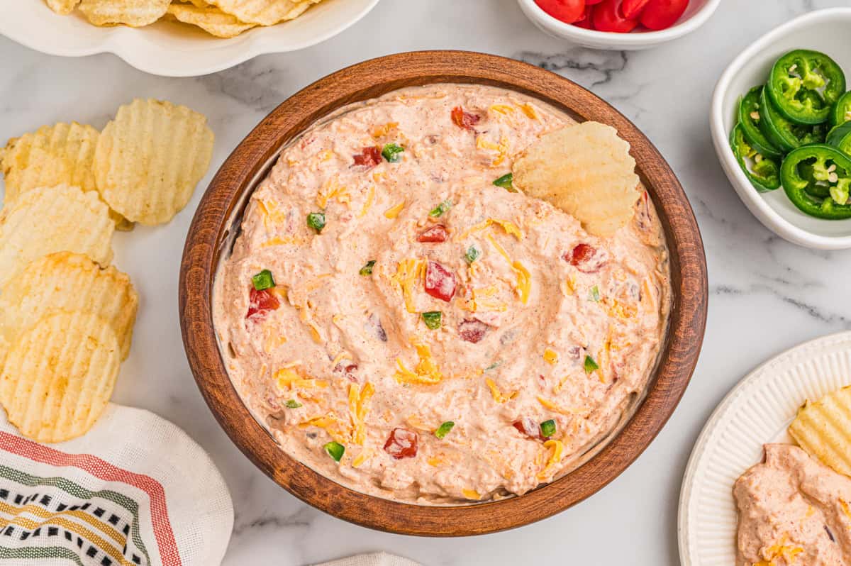 Boat dip in a wooden bowl surrounded by ruffled potato chips, sliced jalapenos, and diced tomatoes.