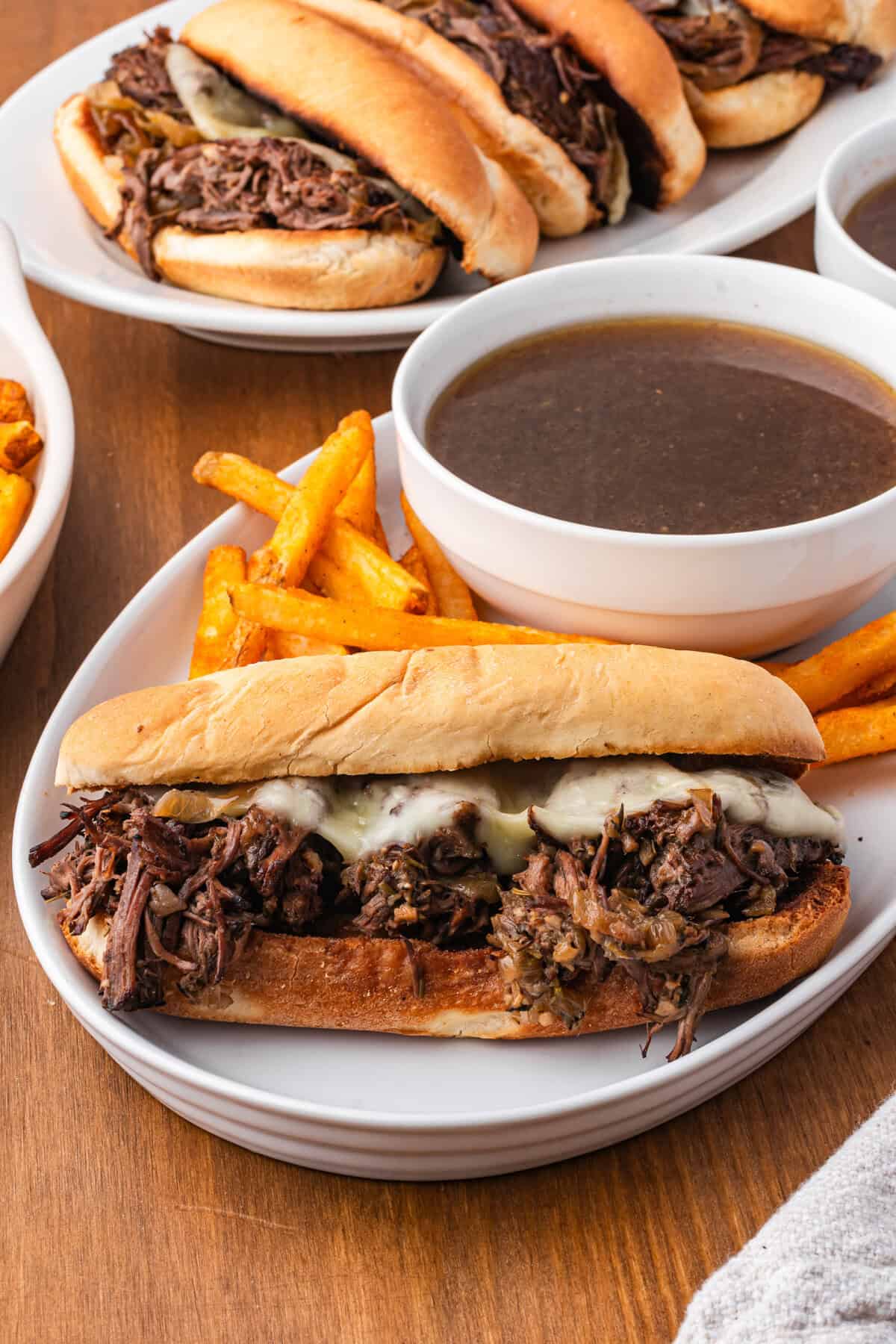 A French dip sandwich plated next to a small bowl of au jus and fries, with more sandwiches on a plate behind.