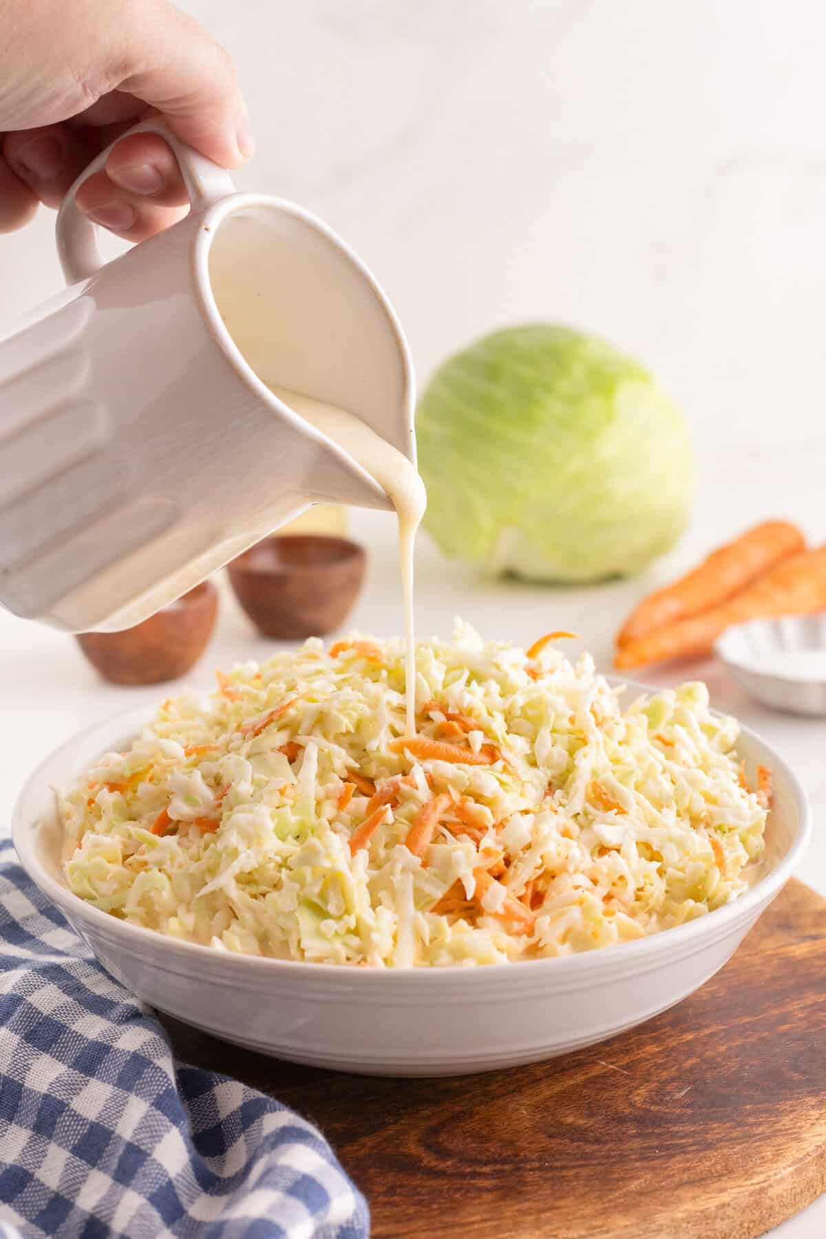 Creamy dressing being poured from a ceramic pitcher onto shredded cabbage and carrots in a white bowl, with a head of cabbage and two carrots in the background.